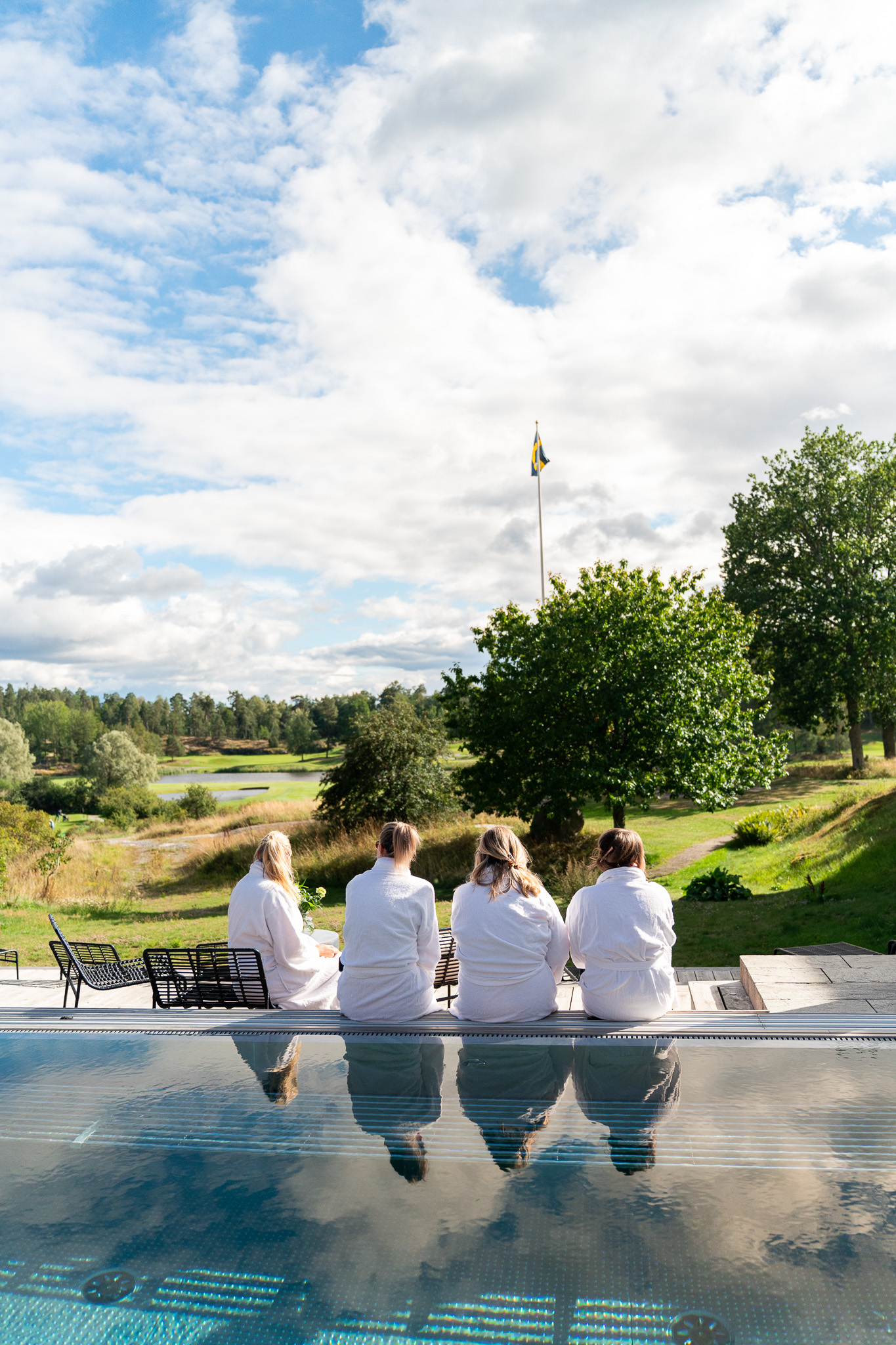 Fyra personer sitter i morgonrockar vid en utomhuspool, de blickar mot grönskande omgivningar