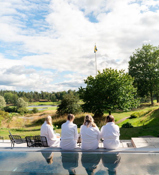 Fyra personer sitter i morgonrockar vid en utomhuspool, de blickar mot grönskande omgivningar